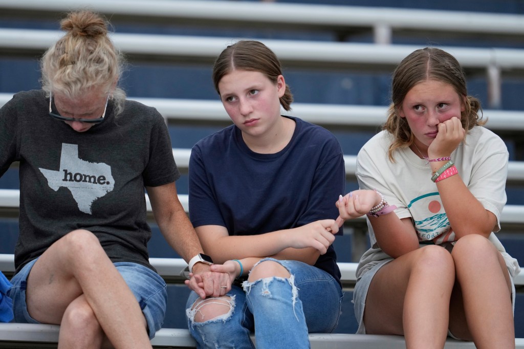 Three women sitting in stadium bleachers, holding hands, appearing somber.