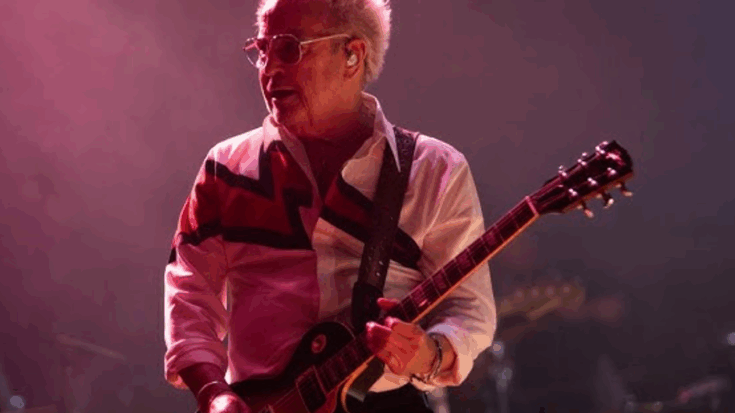 Mick Jones performing on stage with a guitar, wearing a white shirt with bold red and black patterns, under dramatic stage lighting.